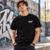 Fiatal férfi fekete, „nyugi” feliratos streetwear pólóban, kis fehér kézírásos mintával a bal mellkason, graffitis városi környezetben.