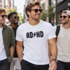 ADHD póló férfi modellen, fehér pólón fekete AD és HD betűkkel, középen villámmal, városi utcán sétáló baráti társaságban.